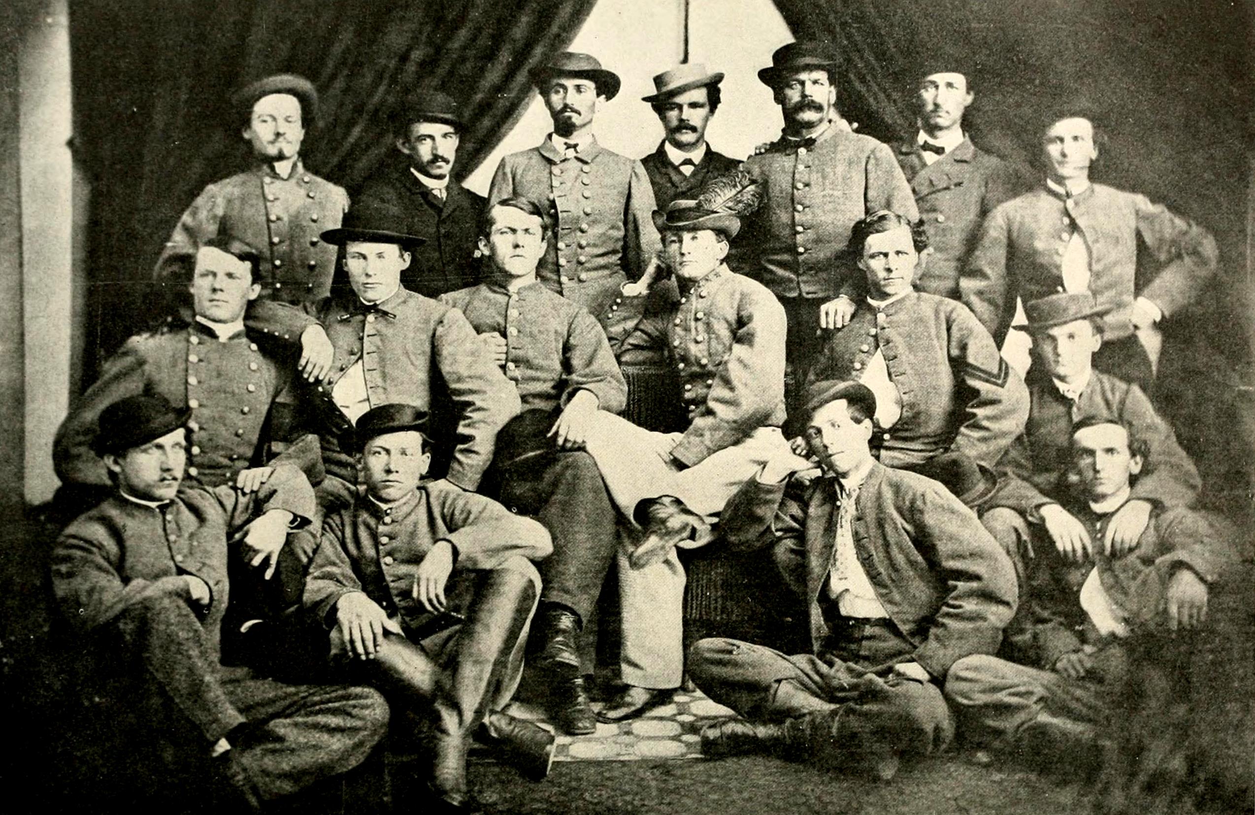 Black-and-white studio group portrait of approximately sixteen white male soldiers in Confederate Army uniforms arranged in three rows — some standing, some seated, and some sitting on the floor. Several wear wide-brimmed hats and double-breasted gray jackets with visible buttons. A draped curtain serves as the backdrop. Civil War era photograph.