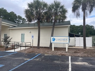 Exterior of the Lakeside at Blue Heron facility, a single-story white building with a sign reading 'Lakeside at Blue Heron' near the entrance. An ADA-accessible ramp with handrails leads to the front door. Several tall sabal palm trees are planted along the front of the building, with a paved parking area in the foreground under an overcast sky.
