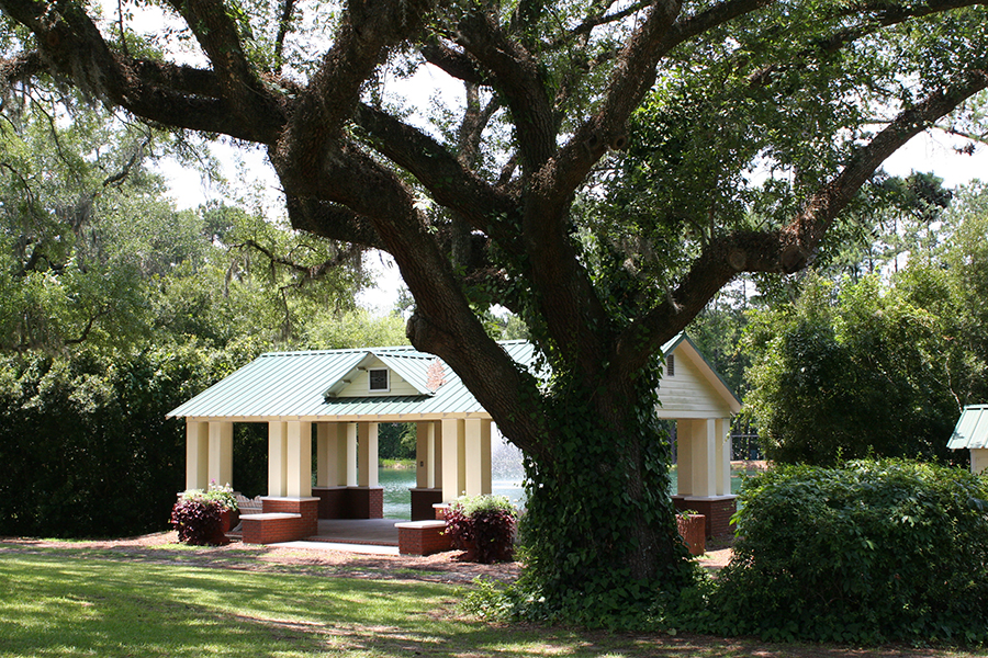 A open-air park pavilion with a green metal roof and cream-colored columns sits beside a pond with a fountain, partially framed by a large sprawling live oak tree draped with Spanish moss. A smaller white building with a matching green roof stands to the right. Lush greenery, trimmed shrubs, and a manicured lawn surround the structures on a sunny day.