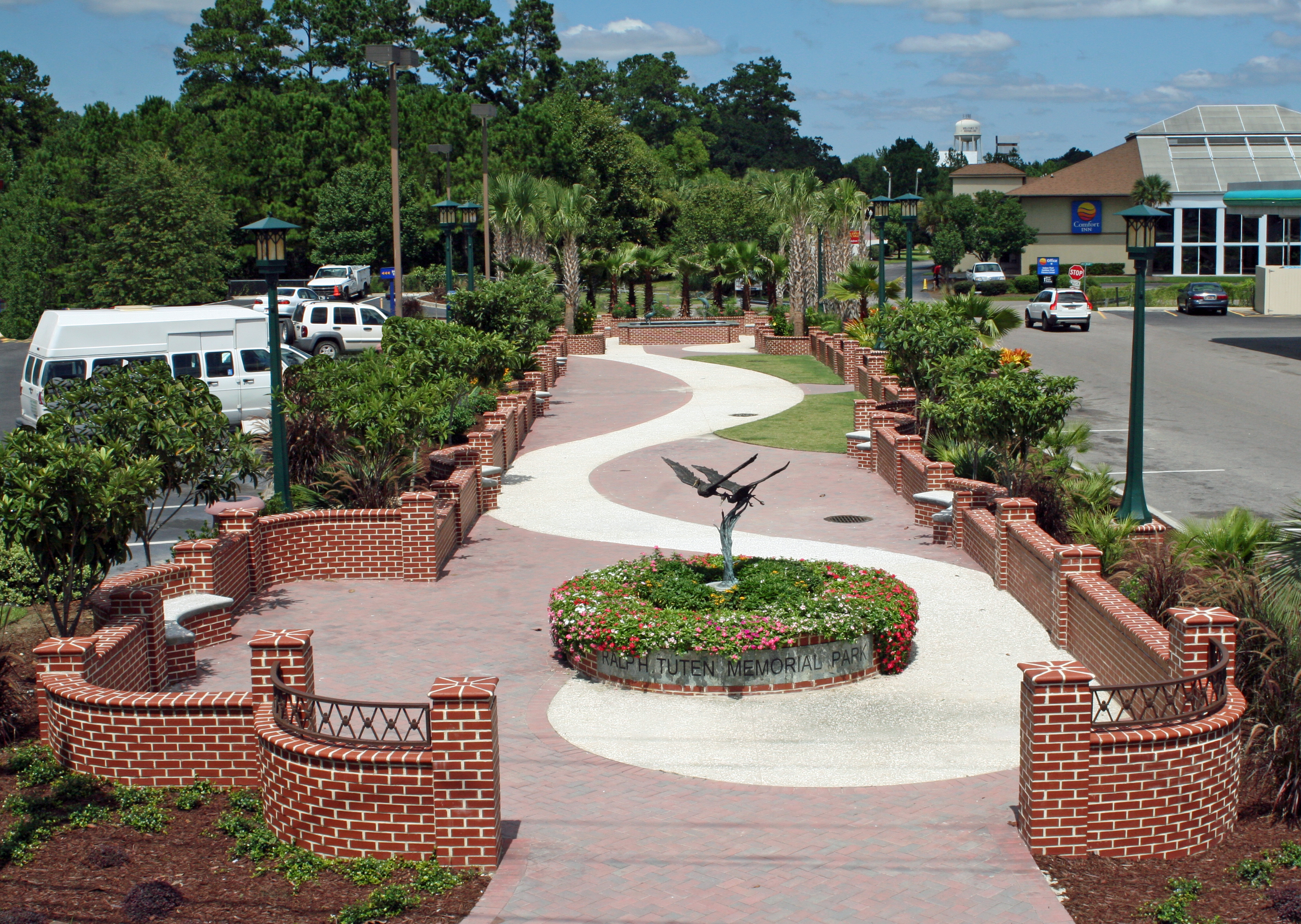 Elevated view of Ralph Tuten Memorial Park showing a wide serpentine brick and paver walkway flanked by low brick walls, ornamental lamp posts, palm trees, and lush landscaping. A circular planter with a bronze heron sculpture and colorful flowers sits at the center of the park with a sign reading 'Ralph Tuten Memorial Park.' A Comfort Inn and parking areas with vehicles are visible on either side, with pine trees and a water tower in the background under a partly cloudy sky.