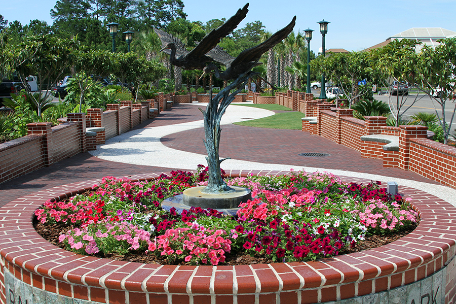 A bronze sculpture of a great blue heron with wings spread wide rises from the center of a circular brick planter overflowing with pink, red, and white petunias at Ralph Tuten Memorial Park. A serpentine brick and paver walkway winds through the landscaped park behind the sculpture, flanked by low brick walls, green lamp posts, palm trees, and pine trees under a clear blue sky.