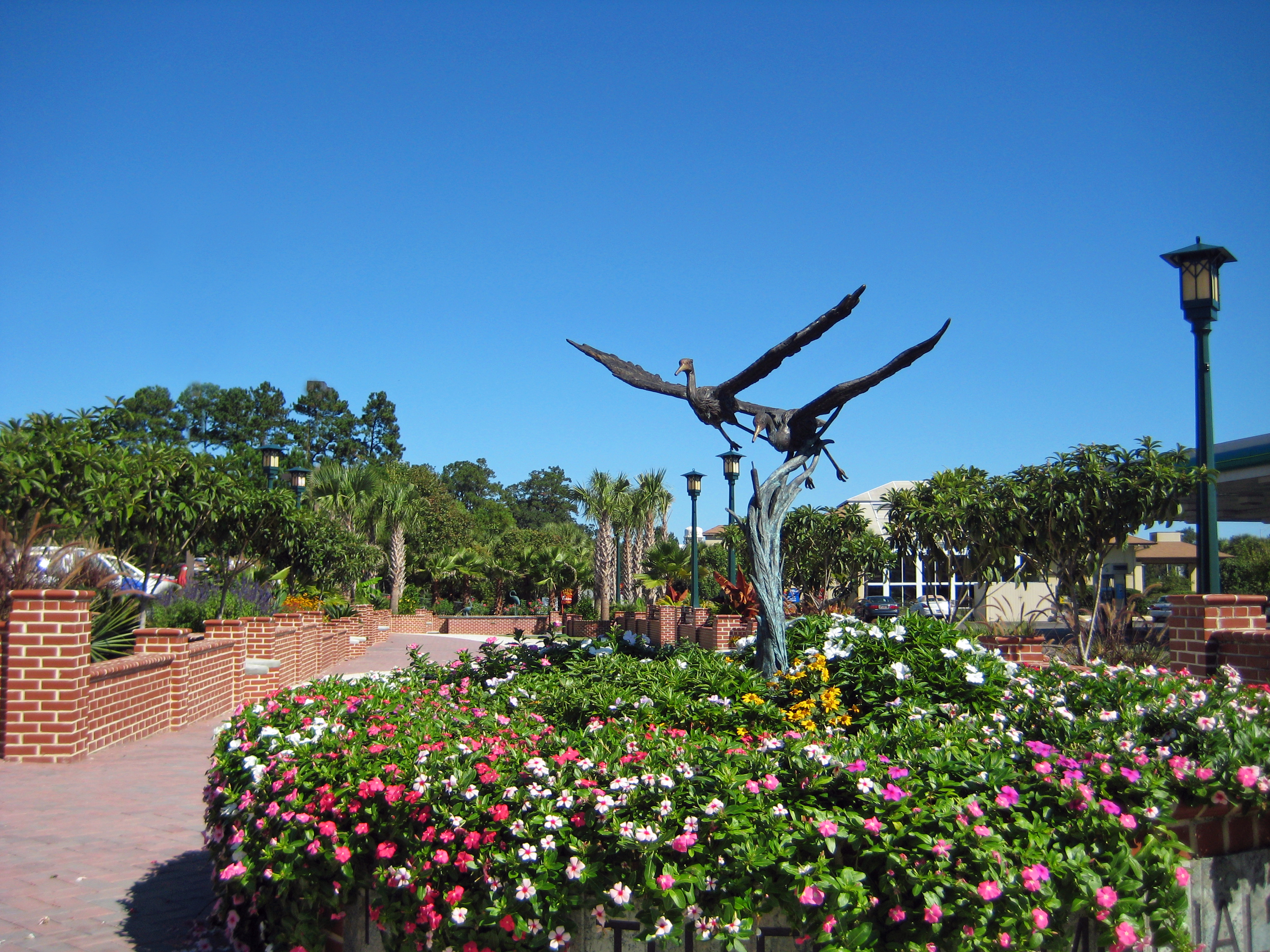 Close-up view of a bronze sculpture depicting two great blue herons in flight, mounted on a twisted branch-like pedestal, rising above a lush circular garden bed filled with pink, white, and yellow flowers at Ralph Tuten Memorial Park. Green lamp posts, low brick walls, palm trees, pine trees, and a commercial building are visible in the background beneath a vivid clear blue sky.