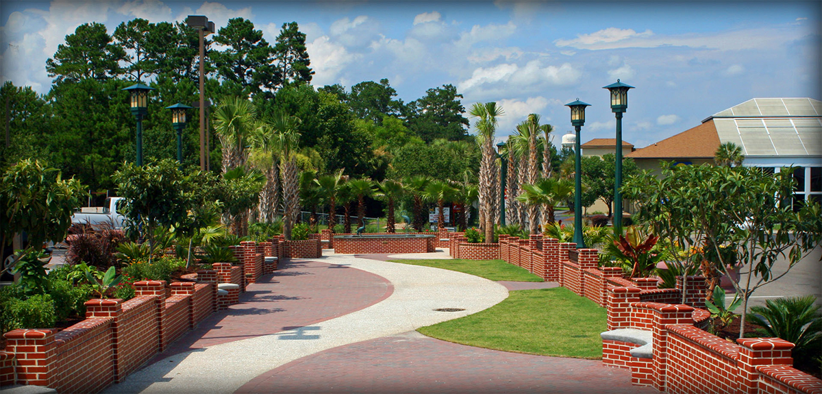 A wide serpentine brick and paver walkway winds through Ralph Tuten Memorial Park, flanked by low red brick retaining walls filled with lush tropical plantings, green grass, and ornamental shrubs. Rows of sabal palm trees and tall green lamp posts line the path, with pine trees in the background and commercial buildings visible on the right under a bright blue sky with scattered clouds.