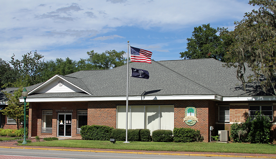 Exterior of the Town of Ridgeland, South Carolina Town Hall, a one-story red brick building located at One Town Square. An American flag and a South Carolina state flag fly from a flagpole in front of the building. The Town of Ridgeland seal is mounted on the facade. Mature oak trees draped with Spanish moss frame the building on a partly cloudy day.