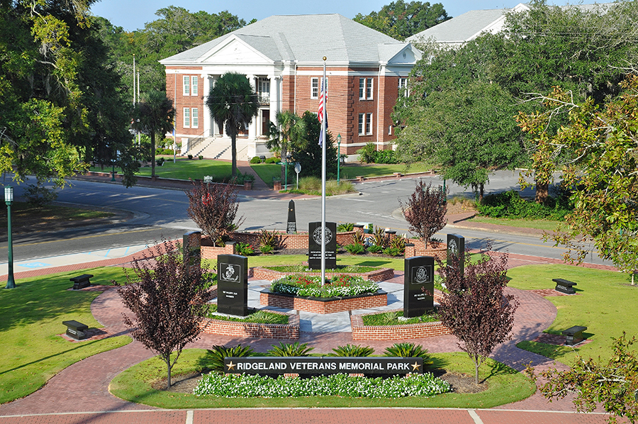 Elevated view of Ridgeland Veterans Memorial Park featuring a circular brick plaza with a central flagpole, military branch memorial monuments, landscaped flower beds, and ornamental trees. A sign at the front reads 'Ridgeland Veterans Memorial Park' with gold stars. A two-story red brick courthouse with white columns stands directly behind the park across the street, framed by mature trees on a clear sunny day.