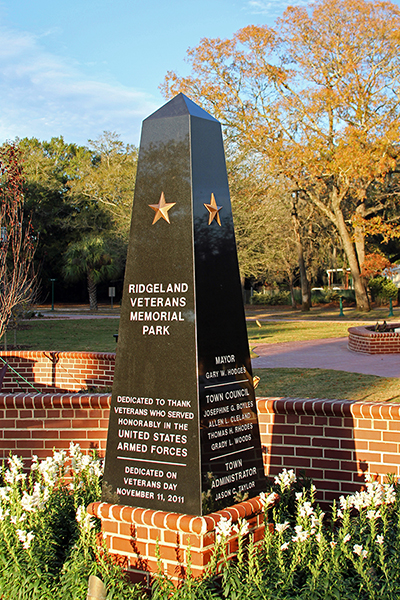 A tall black granite obelisk monument in Ridgeland Veterans Memorial Park bearing two gold stars and engraved white text. The left face reads 'Ridgeland Veterans Memorial Park — Dedicated to thank veterans who served honorably in the United States Armed Forces — Dedicated on Veterans Day, November 11, 2011.' The right face lists the Mayor Gary W. Hodges, Town Council members Josephine G. Bowles, Aileen J. Cleveland, Thomas S.H. Rhodes, and Grady L. Woods, and Town Administrator Jason C. Taylor. The monument sits on a brick base surrounded by white flowers and autumn foliage in the background.