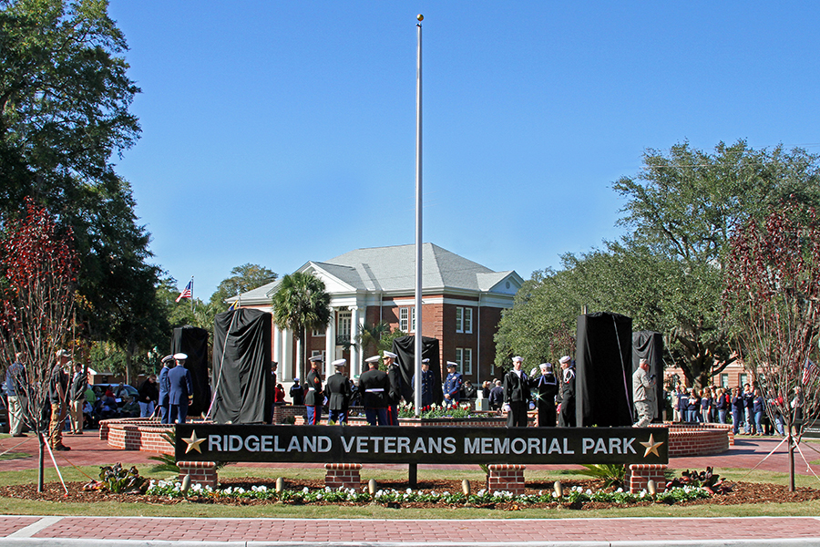 Dedication ceremony at Ridgeland Veterans Memorial Park on a clear sunny day. Members of multiple military branches in dress uniforms stand in formation in the circular brick plaza as black fabric coverings are being removed from the granite memorial monuments. A crowd of onlookers lines both sides of the park. The entrance sign reads 'Ridgeland Veterans Memorial Park' with gold stars. A red brick courthouse with white columns is visible in the background beneath a bright blue sky.
