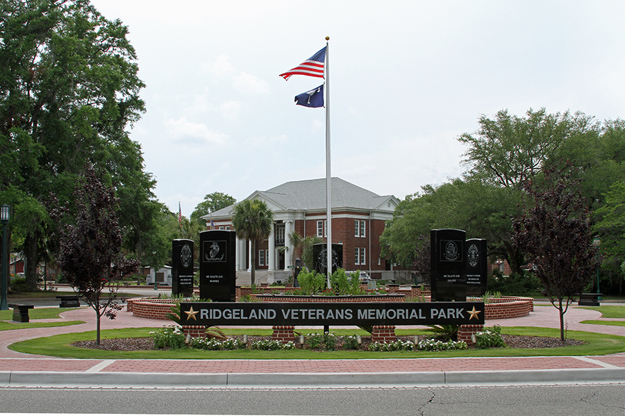 Ground-level view of Ridgeland Veterans Memorial Park showing the entrance sign with gold stars reading 'Ridgeland Veterans Memorial Park,' flanked by black granite military branch memorial monuments and ornamental trees in a circular brick plaza. An American flag and a South Carolina state flag fly from a central flagpole. A red brick courthouse with white columns is visible in the background beneath an overcast sky.