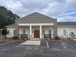 Exterior of a small one-story brick building with a white columned front porch, a brown wooden entry door, decorative iron railings, and white siding on the sides. The building is fronted by a paved parking lot with marked spaces. Trees are visible in the background under an overcast sky.