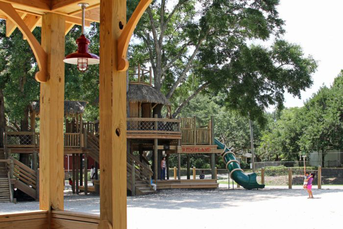 A large multi-level wooden treehouse playground structure with a thatched roof, walkways, and a green tube slide, labeled 'Ridgeland,' surrounded by mature oak trees. Two children are visible near the slide on a gravel play surface. The photo is taken from beneath a nearby wooden pavilion with a red pendant light in the foreground.